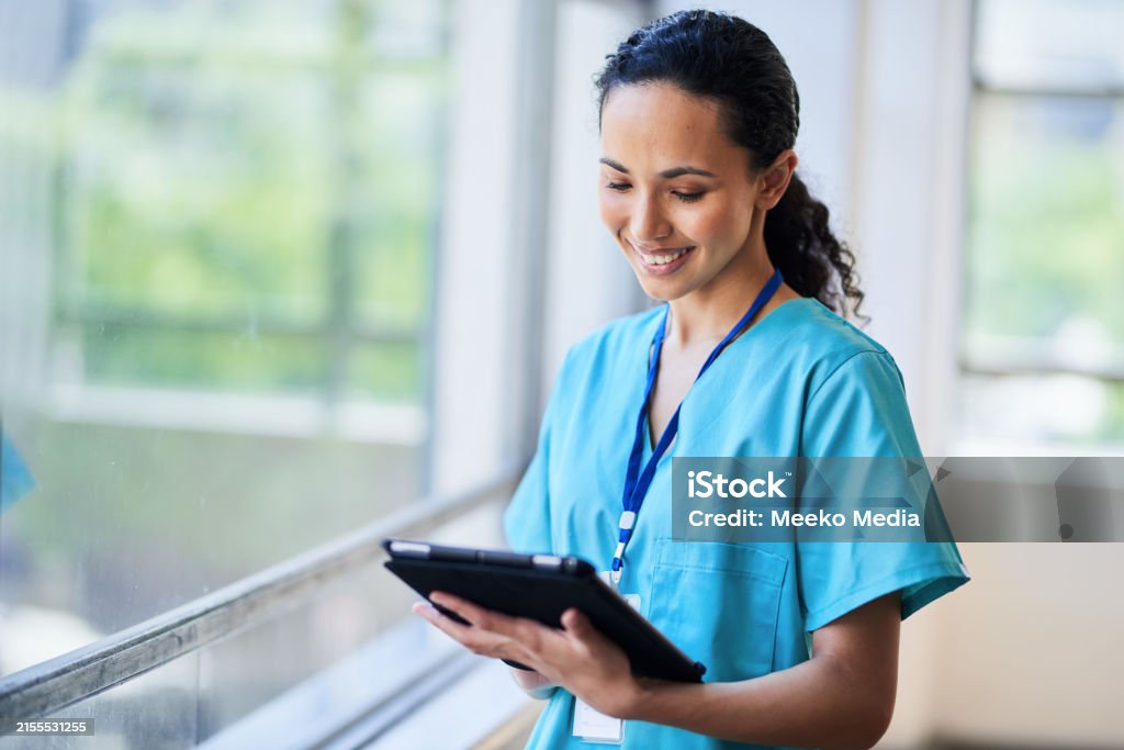 Smiling Nurse in Hospital Scrubs Using Digital Tablet in Modern Medical Facility Smiling nurse wearing hospital scrubs and holding a digital tablet while standing in a modern medical facility. The image portrays a professional medical worker in a healthcare environment, using technology for patient care. Digital Tablet Stock Photo