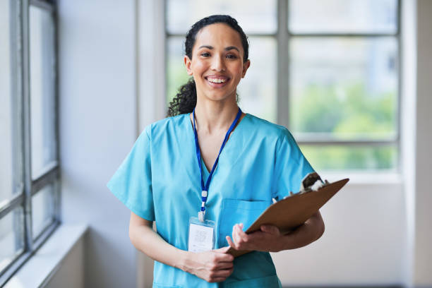 Smiling Medical Professional in Scrubs Holding a Clipboard in Hospital Setting Portrait of a cheerful medical professional in blue scrubs holding a clipboard. The multi-ethnic healthcare worker is standing in a bright hospital hallway, symbolizing diversity, dedication, and expertise in the medical field. nurse stock pictures, royalty-free photos & images