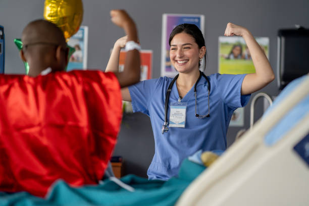 Bravely Waiting Through Chemo A sweet little boy of African decent, who is battling Cancer, sits in his hospital bed with a superhero cape on, as he patiently waits for his Chemotherapy treatment to run. He is flexing his muscles as she shows his nurse how strong he is! red medical scrubs stock pictures, royalty-free photos & images
