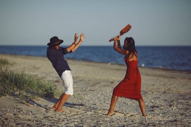 Woman attacking a man with a guitar on a sandy beach. Crisis of relationships during vacation on the beach at sunset, with the woman in a red dress and the man in a hat and shorts.
