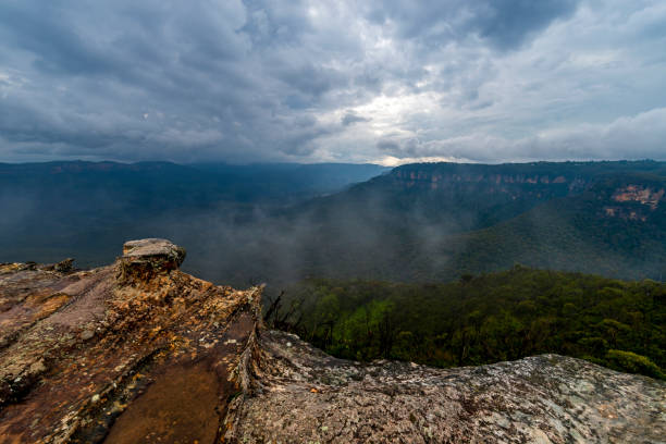View of deep valley and rolling hills from a rocky cliff edge. Gloomy day with dramatic cloudscape. White mist rising from the valley trough (mid-ground).