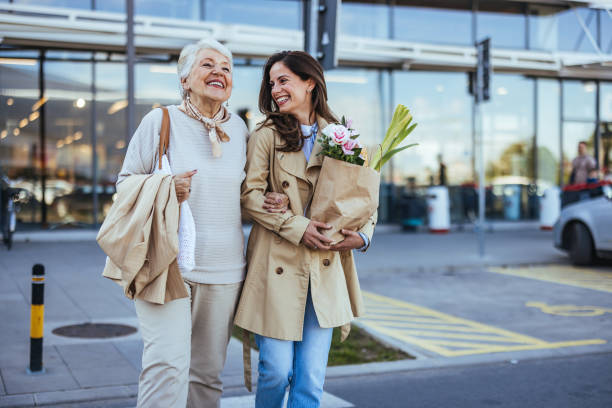 Family Bonding Over Shopping Errands A joyful adult daughter walks with her elderly mother outside a store, both carrying grocery bags and sharing a light-hearted moment together. mall walker stock pictures, royalty-free photos & images
