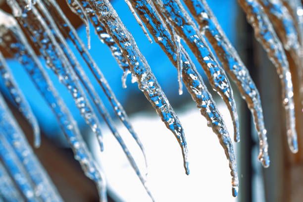 Icicle against light blue sky. Drop of melted snow falls down. Closeup. Illustration about end of winter or beginning of spring.