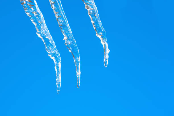 Icicle against light blue sky. Drop of melted snow falls down. Closeup. Illustration about end of winter or beginning of spring. Thaw. Macro image. Copy space.
