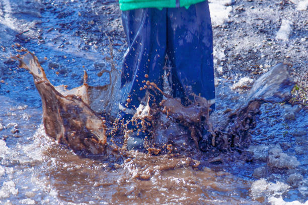 Child in rubber boots and protective rubber pants jumping in the muddy puddle