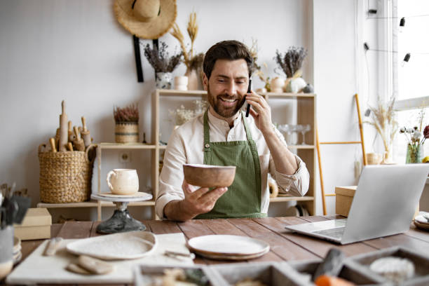 istock Young bearded man potter taking order for purchase of tableware. 2155589080