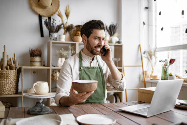 istock Young bearded man potter taking order for purchase of tableware. 2155589094
