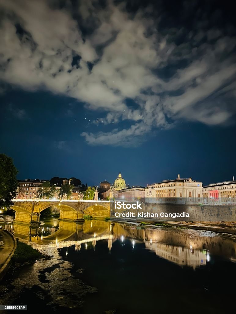 City lights reflecting off River Tiber, looking towards St Peter’s dome in Rome. View of Ponte Vittorio Emanuele II bridge and St Peter’s Basilica with city lights beautifully glowing in Rome. Night sky with a cloudscape. Cityscape Stock Photo City lights reflecting off River Tiber, looking towards St Peter’s dome in Rome. View of Ponte Vittorio Emanuele II bridge and St Peter’s Basilica with city lights beautifully glowing in Rome. Night sky with a cloudscape. Cityscape Stock Photo