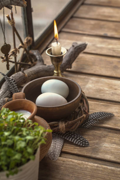 Atmospheric spring altar with burning candle, bowl with eggs, the first greens in the pot and birds feathers on wooden surface, vertical image, selected focus. Ostara, Imbolc, pagan, religion, magic.