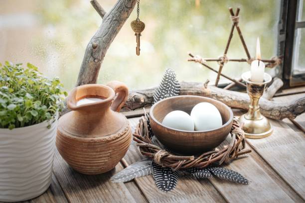 istock Home atmospheric spring altar with willow wreath, clay jug with milk, bowl with eggs, the first greens in the pot, candle and birds feathers on wooden surface, selected focus. 2155605208