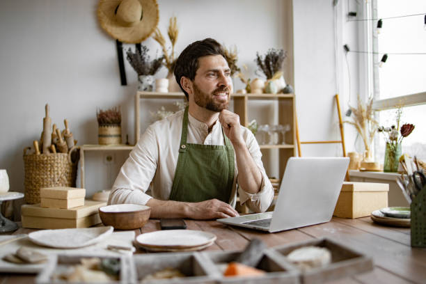 istock Relaxing man in apron enjoys break while sitting at table in pottery shop. 2155605396
