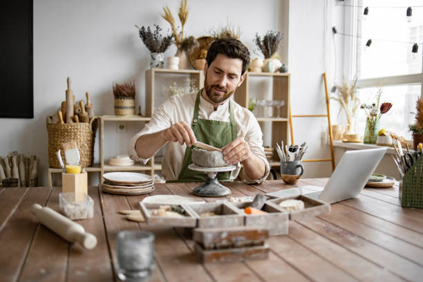istock Bearded male seated at wooden table with various tools, materials, and laptop. 2155607513