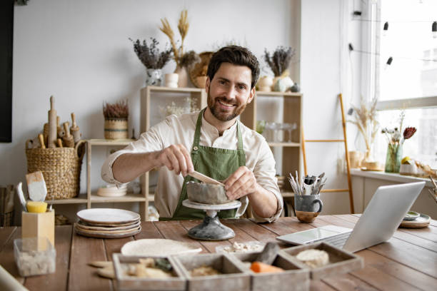 istock Bearded male seated at wooden table with various tools, materials, and laptop. 2155607515