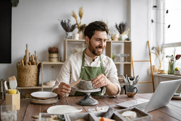 istock Bearded male seated at wooden table with various tools, materials, and laptop. 2155607516