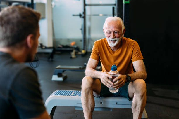 Elderly man sitting on a bench holding a water bottle, smiling after a workout session in a gym with a personal trainer.