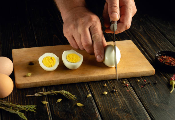 istock Hands with a knife cut an egg. Sliced eggs on a kitchen board for preparing a diet lunch with aromatic spices. Advertising space 2155627650