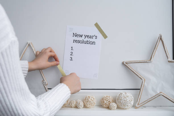 Unrecognizable woman creating vision board. NEW YEAR'S RESOLUTION checklist text on paper note stick on while wall with cozy minimalistic handmade Christmas decor. New year aims resolutions. Low key festive Planning and setting goals concept mujer irreconocible creando tablero de visión. resoluciÓn de aÑo nuevo texto de la lista de verificación en papel nota adhesiva mientras se pega en la pared con una acogedora decoración navideña minimalista hecha a mano. propósitos de año nuevo. pl