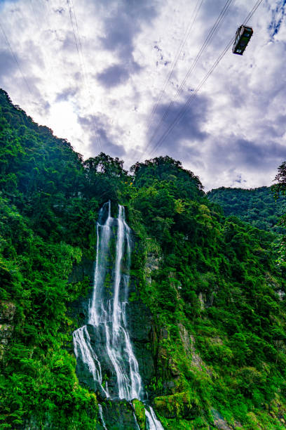 A waterfall is shown in the foreground of a lush green mountain. The waterfall is surrounded by trees and is a beautiful sight to behold. The sky is cloudy, adding to the moody atmosphere of the scene