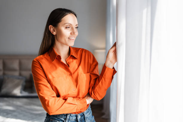istock Smiling woman wearing orange shirt , looking at window while standing at home 2155681609