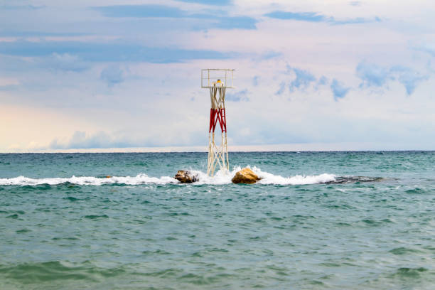 A small, minimalist lighthouse stands on rocky terrain amidst the waves in the Aegean Sea, Greece. The lighthouse, with its red and white structure, contrasts against the calm blue-green waters and a slightly cloudy sky, symbolizing navigation and safety.