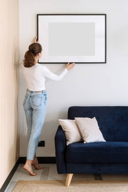 Woman decorated cozy home with art work in picture frame Full length view of woman in casual wear decorated cozy home with art work in picture frame. Copy space poster in living room over comfortable couch. Female install placard on wall, back rear view hanging art on wall stock pictures, royalty-free photos & images