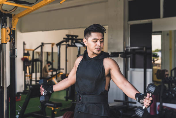 istock A tanned and handsome asian guy doing standing dumbbell scaption raises midway at the gym. Wearing a low cut tank top. Weight training at the gym. 2155703068