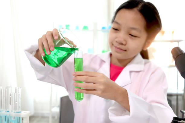 Hand of adorable Asian schoolgirl in lab coat pouring green reagent solution from beaker into test tube, doing science experiments, happy smiling scientist kid having fun during doing experiments, child using microscope, apparatus and lab equipment for research. Science education for kid concept. hand of adorable asian schoolgirl in lab coat pouring green reagent solution from beaker into test tube, doing science experiments, happy smiling scientist kid having fun during doing experiments, child using microscope, apparatus and lab equipment for re