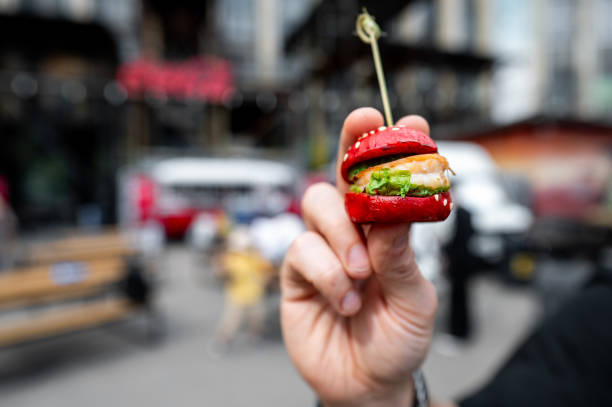 istock A miniature burger with a sesame seed bun, lettuce, and tomato held between fingers against a blurred urban background. 2155723154