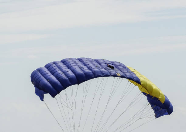 A solitary skydiver descends under a bright, multicolored canopy, approaching a distant landing area. The vast blue sky and open space emphasize freedom, adventure, and precision in flight. The scene captures the thrill of air sports, control in descent, and the calm before a safe landing.