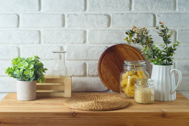 istock Empty wooden kitchen table with wicker place mat, food jars and plants over white brick wall  background.  Kitchen mock up for design and product display. 2155742102