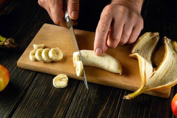 istock Finely sliced banana on a kitchen wooden board. Cooking a fruit dish on the kitchen table with the hands of a chef. Place for advertising 2155746915