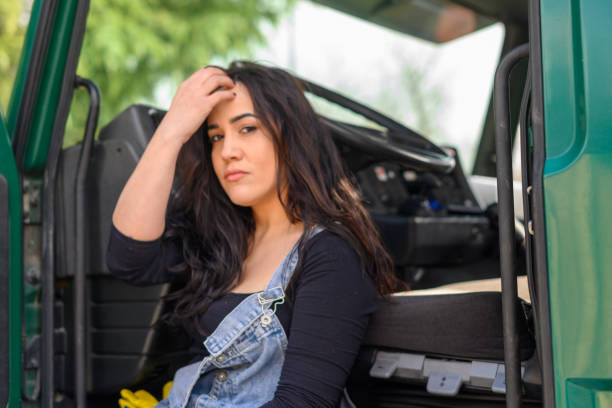 Hispanic young model poses in black shirt and blue overalls lean up set a big green cargo truck in construction site.