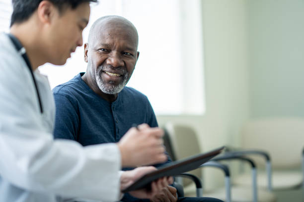 Senior Medical Exam A senior gentleman of African decent sits in a waiting room beside his doctor as they review test results on a tablet together. patient stock pictures, royalty-free photos & images