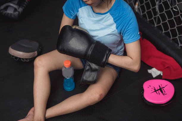 A novice female MMA practitioner removes her boxing gloves while sitting down on the mat, exhausted.