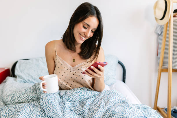 istock Young beautiful woman using mobile phone while drinking coffee in the bed 2155812083