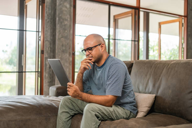 istock Young asian man sitting on sofa looking at display screen tablet feeling serious stressed. He using texting on tablet reading bad news today in the morning 2155817040
