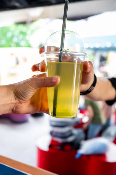 istock Close-up of two hands exchanging a plastic cup with a yellow-green beverage and a straw against a blurred outdoor background 2155820057
