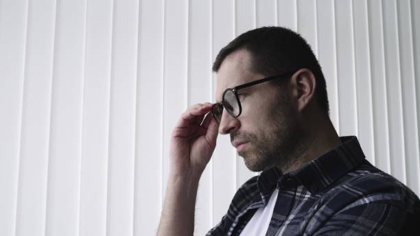 Thoughtful bearded young man standing near window and taking off his glasses. Lifestyle concept. Side view. Slow motion. Copy space