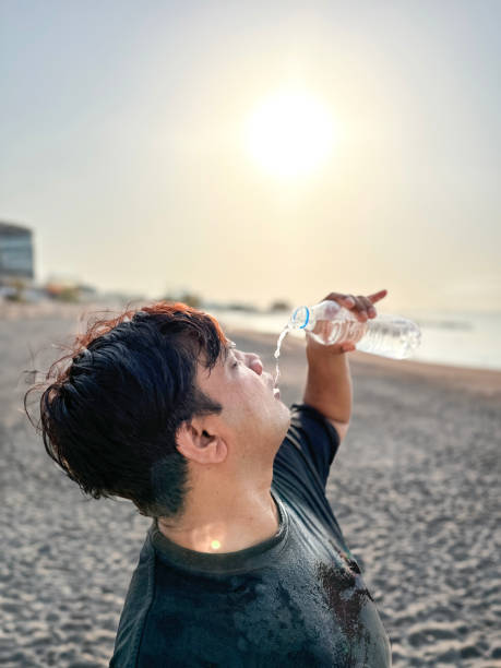 Stock photo of Indian man drenched in sweat drinking cold water from clear plastic bottle on sandy beach to cool himself down from scorching sun , heatwave with temperatures as high as 40c and above, full sun, hydrating in hot warm weather