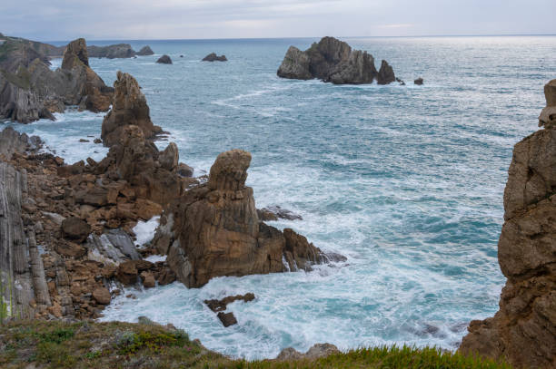 A rugged coastline with jagged rocks protruding from the sea, grass-covered cliffs, and a cloudy sky. The naturalistic style captures the wild beauty of the coastal landscape, suitable for themes of travel, nature conservation, or outdoor adventure. The w