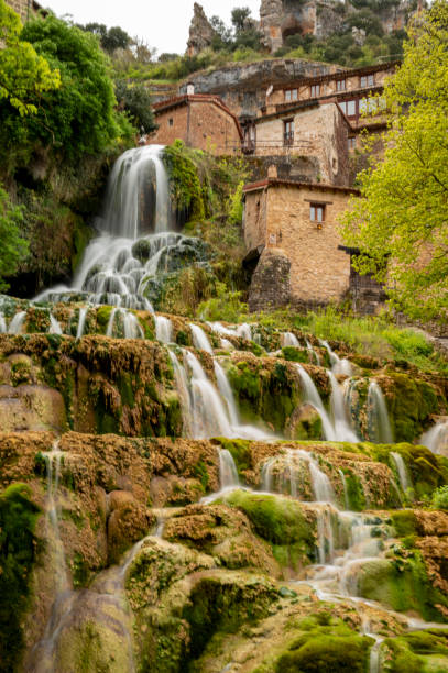 The image portrays a multi-tiered waterfall cascading over moss-laden rocks with a traditional stone edifice nestled against a verdant hillside. The nature photography style captures the essence of the environment, suitable for design and print with a focus on natural landscapes. The vertical composition enhances the images depth and the flow of the waterfall. la imagen retrata una cascada de varios niveles que cae en cascada sobre rocas cargadas de musgo con un edificio de piedra tradicional ubicado contra una ladera verde. el estilo de fotografía de naturaleza captura la esencia del entorno, adecuado para el