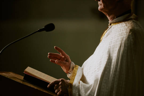Side view crop shot of senior Caucasian Catholic priest standing at lectern with Bible book on it preaching sermon in Church