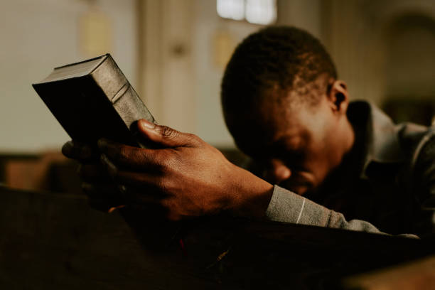 Young Black Parishioner With Bible In Hands Selective focus shot of African American man sitting on bench in Catholic church holding Bible book in hands black gospel church stock pictures, royalty-free photos & images