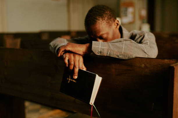 Unrecognizable Black Man Praying In Church Selective focus shot of unrecognizable African American man sitting on pew in Catholic church holding Bible book and praying to God black gospel church stock pictures, royalty-free photos & images