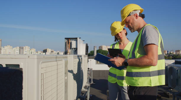 istock Maintenance workers or engineers wearing protective helmets and visibility yellow vests examining a new AC unit fan on the roof and taking notes 2155840385