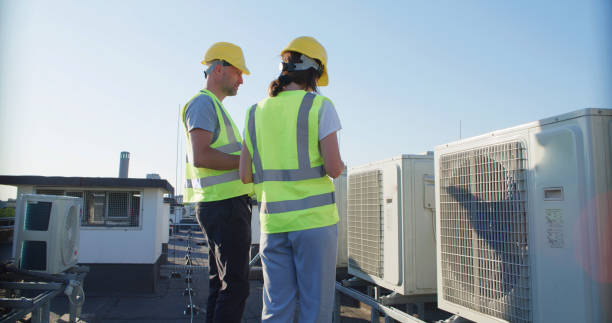 istock Maintenance workers or engineers wearing protective helmets and yellow vests examining a new AC unit fan on the roof outdoors and taking notes 2155840396