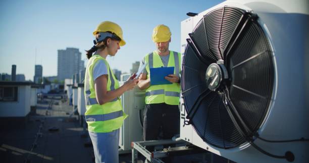 istock Maintenance workers or engineers wearing helmets and visibility yellow vests examining a new AC unit fan on the roof outdoors together and taking notes 2155840453