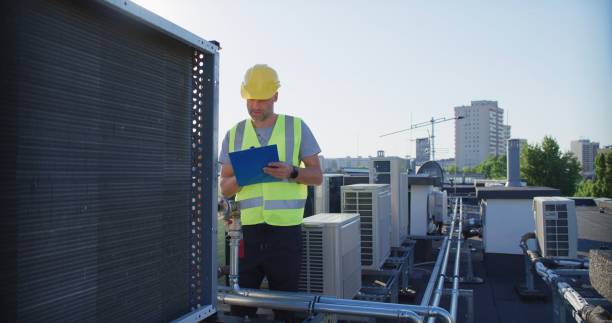istock Maintenance worker wearing protective helmet and visibility yellow vest examining AC unit fan outdoors and taking notes 2155840492