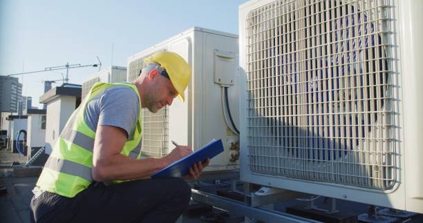 istock Maintenance worker or engineer wearing protective helmet and visibility yellow vest examining a new AC unit fan on the roof outdoors and taking notes 2155840502