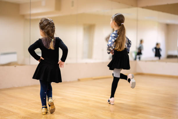 Two young girls practicing dance moves in a dance studio with mirrors. They are wearing dance attire and focusing on their reflection.